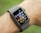 A smartwatch with a black screen and multiple colorful app icons is worn on a person's wrist. The watch band is gray, and the image background is blurred green, suggesting an outdoor setting.