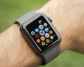A smartwatch with a black screen and multiple colorful app icons is worn on a person's wrist. The watch band is gray, and the image background is blurred green, suggesting an outdoor setting.