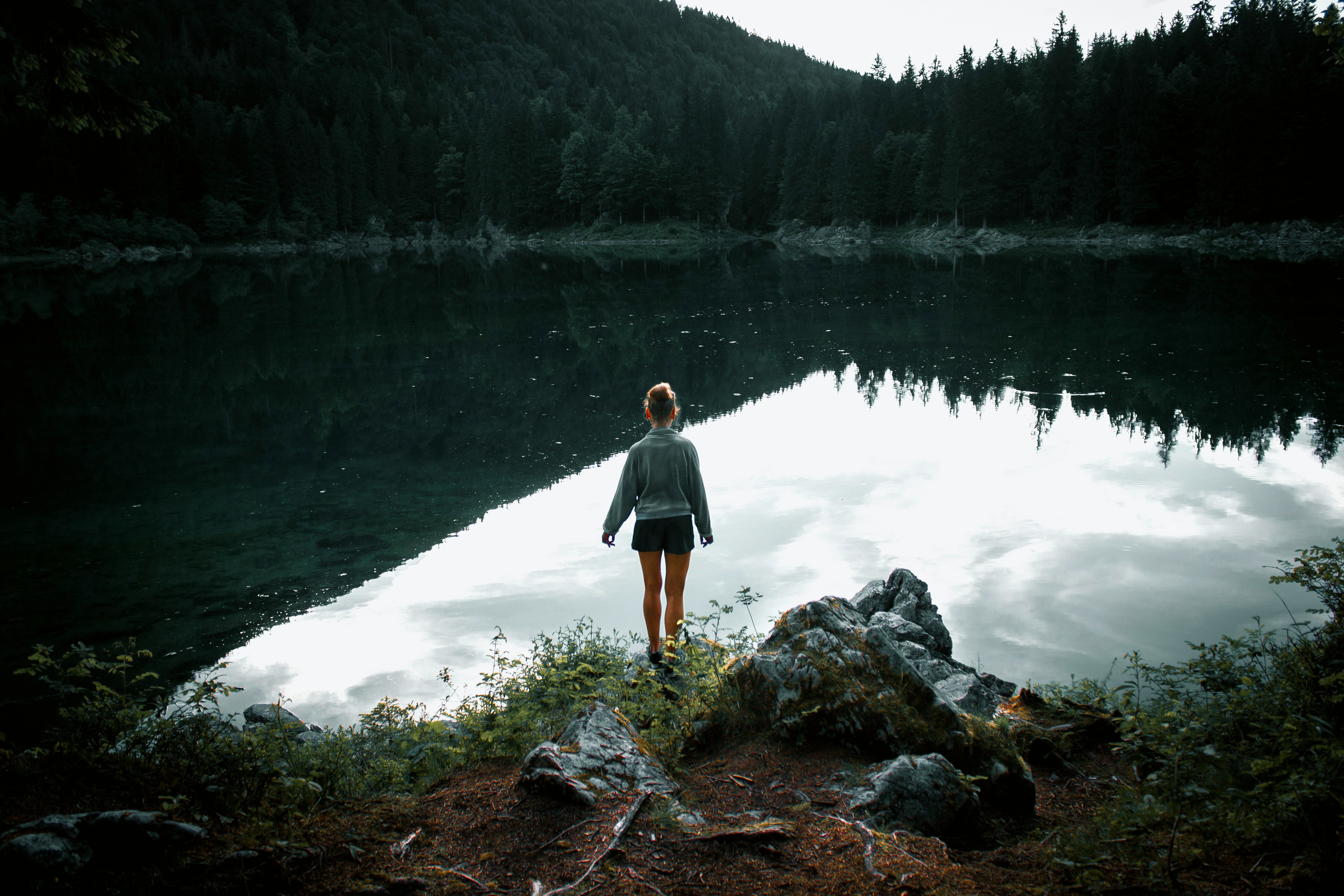 woman standing on rock near body of water facing trees during daytime