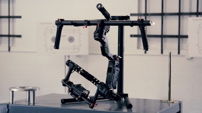 A professional camera stabilizer setup is displayed on a metallic surface against an industrial-style backdrop. The equipment consists of black components with multiple joints, handles, and electronic parts, indicating it is designed for filming and shooting stability.