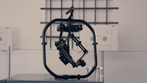 A black, mechanical device with a central structure enclosed in a circular frame is set against a gray industrial background. The device appears complex with multiple joints and components, possibly for engineering or robotics applications.