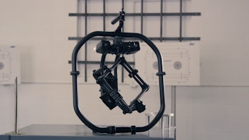 A black, mechanical device with a central structure enclosed in a circular frame is set against a gray industrial background. The device appears complex with multiple joints and components, possibly for engineering or robotics applications.