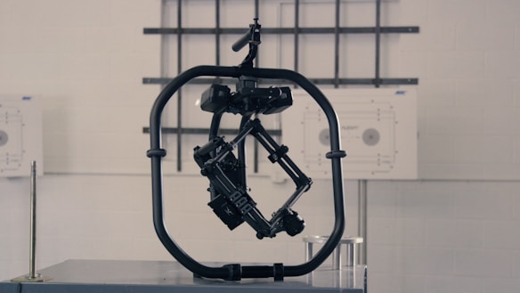 A black, mechanical device with a central structure enclosed in a circular frame is set against a gray industrial background. The device appears complex with multiple joints and components, possibly for engineering or robotics applications.