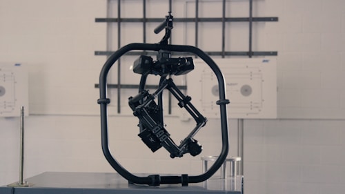 A black, mechanical device with a central structure enclosed in a circular frame is set against a gray industrial background. The device appears complex with multiple joints and components, possibly for engineering or robotics applications.