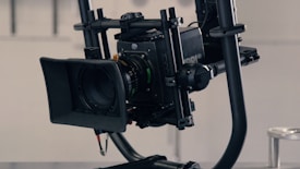 A professional camera mounted on a stabilizing rig. The camera has a large lens with a lens hood attached and intricate controls visible. The rig features carbon fiber rods and joints, indicating a sturdy build suitable for smooth filming.
