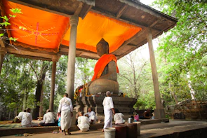 A large Buddha statue sits under a pavilion adorned with a bright orange canopy. Several people in white clothing are gathered around the statue, engaged in various activities including prayer and meditation. The setting is outdoors, surrounded by lush green trees, creating a peaceful and serene atmosphere.