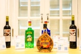 A selection of colorful liquor bottles is displayed on a wooden surface in front of a white window with shutters. There are five bottles in total, each with distinct colors and labels. In the foreground, a prominent bottle labeled 'Kizomba' features an amber hue. Two placards with text and QR codes are placed next to the bottles.