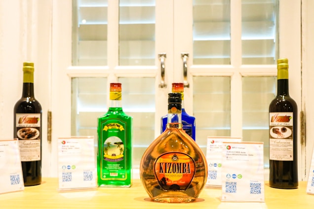 A selection of colorful liquor bottles is displayed on a wooden surface in front of a white window with shutters. There are five bottles in total, each with distinct colors and labels. In the foreground, a prominent bottle labeled 'Kizomba' features an amber hue. Two placards with text and QR codes are placed next to the bottles.
