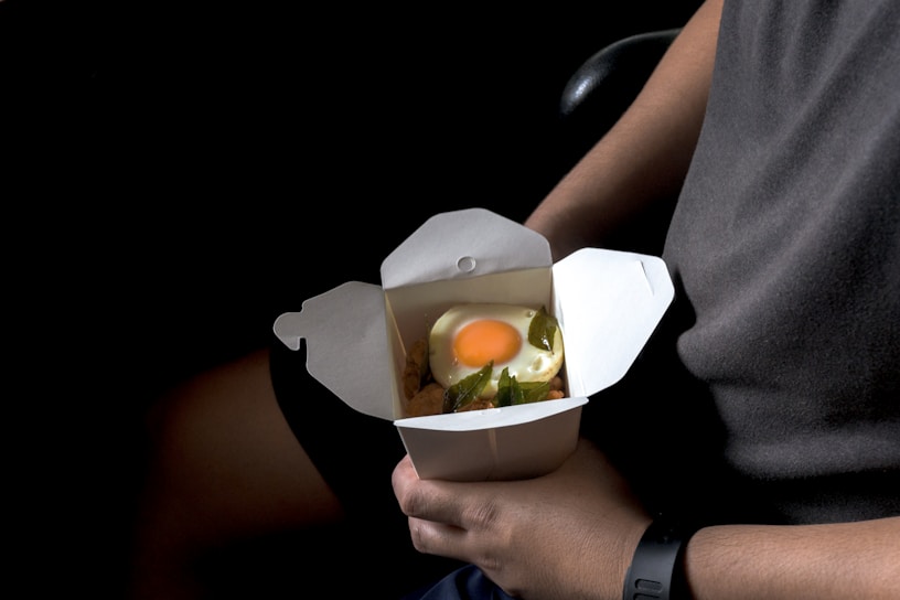A person is holding a white takeout box containing food. Inside the box, there's a dish topped with a sunny side up egg, garnished with green leaves over some other ingredients.
