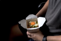 A person is holding a white takeout box containing food. Inside the box, there's a dish topped with a sunny side up egg, garnished with green leaves over some other ingredients.