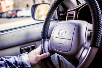 A close-up view of a car's steering wheel with a hand gripping it. The steering wheel displays the Toyota logo, and the dashboard is visible in the background. The interior appears clean and well-maintained with muted lighting.