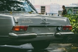 A classic Mercedes-Benz car is parked outdoors, with the model designation 280 SL and the word AUTOMATIC visible on its rear. The car's chrome bumper reflects light, and there are a few leaves scattered on its surface. In the blurred background, a person is partially visible, standing near some greenery with buildings further back.