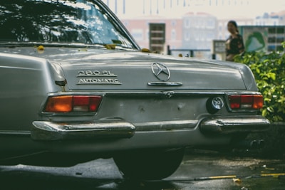 A classic Mercedes-Benz car is parked outdoors, with the model designation 280 SL and the word AUTOMATIC visible on its rear. The car's chrome bumper reflects light, and there are a few leaves scattered on its surface. In the blurred background, a person is partially visible, standing near some greenery with buildings further back.