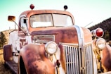 A close-up view of an old rusted truck, featuring a weathered exterior with peeling paint. The headlights are vintage, and the body displays signs of aging. This truck is parked outdoors with a background of trees and a blue sky, suggesting a serene rustic setting.