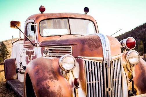 A close-up view of an old rusted truck, featuring a weathered exterior with peeling paint. The headlights are vintage, and the body displays signs of aging. This truck is parked outdoors with a background of trees and a blue sky, suggesting a serene rustic setting.