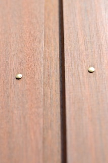 Close-up view of a wooden surface with two metal nails visible. The wood has a natural grain texture and a warm, light brown color.