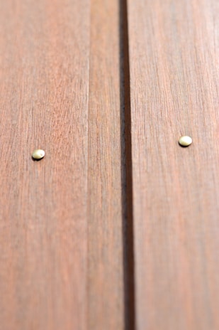Close-up view of a wooden surface with two metal nails visible. The wood has a natural grain texture and a warm, light brown color.
