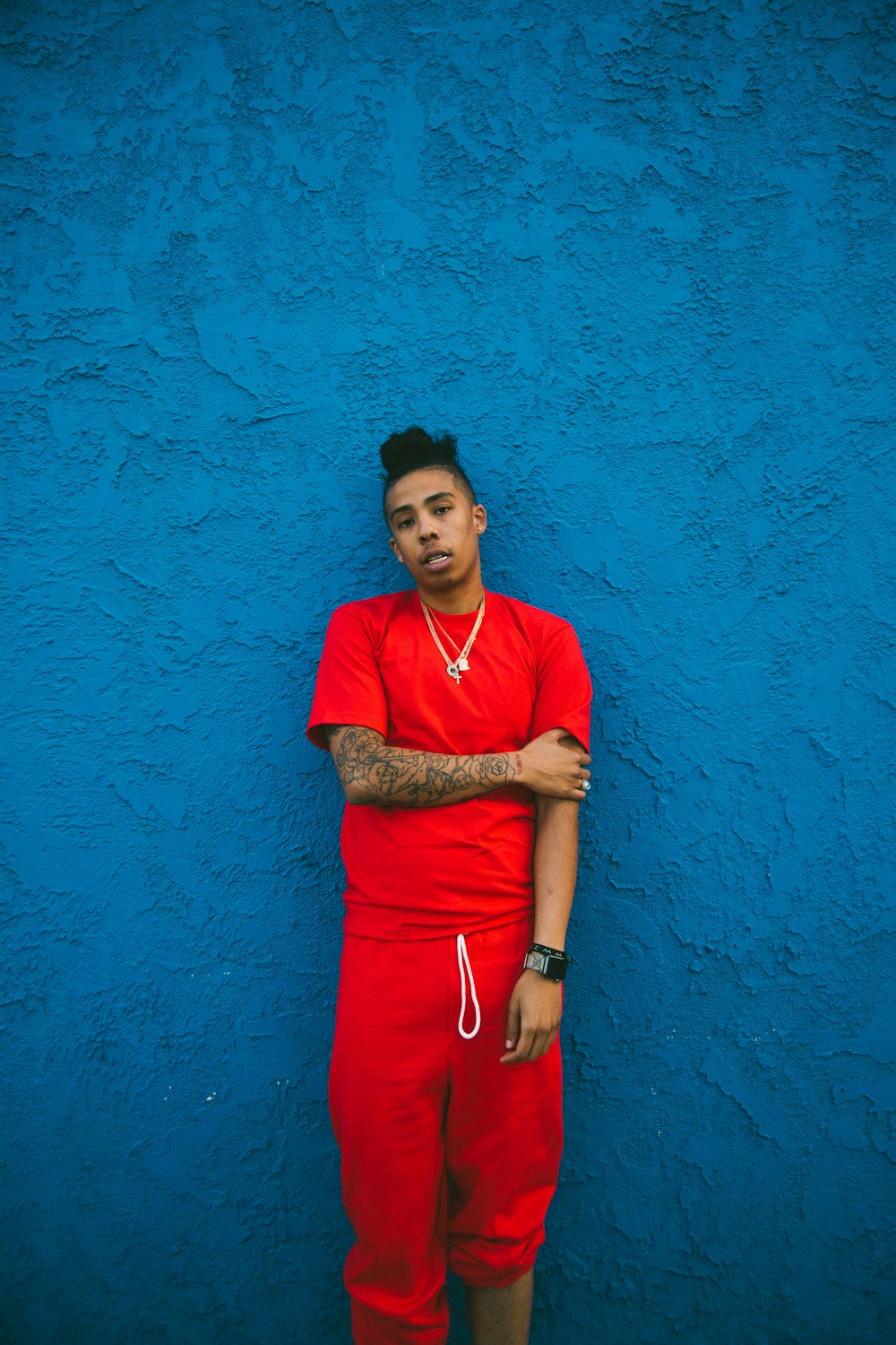 man wearing red crew-neck t-shirt and drawstring pants leaning on blue wall