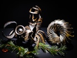 A collection of intricately designed metallic objects resembling machinery parts. These objects feature gears and coil-like structures, arranged artistically against a dark background. Pine branches with green needles add a contrasting natural element at the bottom.