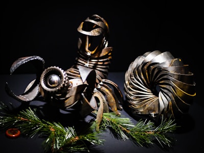 A collection of intricately designed metallic objects resembling machinery parts. These objects feature gears and coil-like structures, arranged artistically against a dark background. Pine branches with green needles add a contrasting natural element at the bottom.