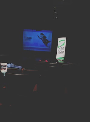 A dimly lit room with a projector screen displaying an animated character and text. To the right, a standing banner with the words 'San Isidro'. Several empty seats with some garments and papers are visible in the foreground.
