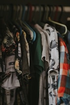 A collection of various t-shirts hangs on a clothes rack, showcasing different patterns and colors. Some shirts have graphic designs, while others feature solid colors or plaid patterns. The hangers are made of plastic, with multiple colors, including blue, green, and brown.