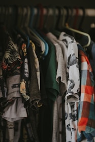 A collection of various t-shirts hangs on a clothes rack, showcasing different patterns and colors. Some shirts have graphic designs, while others feature solid colors or plaid patterns. The hangers are made of plastic, with multiple colors, including blue, green, and brown.