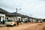 A row of newly constructed single-story houses with dark grey roofs and white walls, along a dirt road. Two utility poles with wires are visible, and there are street lamps attached to the poles. A small truck with a signage and a person standing next to it are in the foreground. Another larger utility truck is visible further down the road. The overall scene appears to be at a construction site or newly developed residential area under development.
