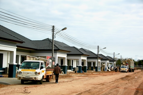 A row of newly constructed single-story houses with dark grey roofs and white walls, along a dirt road. Two utility poles with wires are visible, and there are street lamps attached to the poles. A small truck with a signage and a person standing next to it are in the foreground. Another larger utility truck is visible further down the road. The overall scene appears to be at a construction site or newly developed residential area under development.