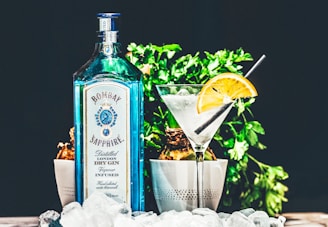 A blue bottle of Bombay Sapphire gin is placed next to a cocktail glass garnished with a slice of orange. The setting includes green plants in the background and ice cubes scattered in the foreground, creating a refreshing and stylish presentation.