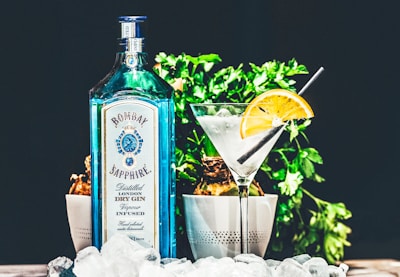 A blue bottle of Bombay Sapphire gin is placed next to a cocktail glass garnished with a slice of orange. The setting includes green plants in the background and ice cubes scattered in the foreground, creating a refreshing and stylish presentation.