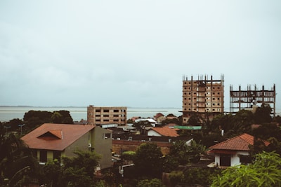 A serene coastal townscape features buildings with red roofs surrounded by lush greenery. Two unfinished high-rise structures stand out against the backdrop of a wide river or lake, under an overcast sky.