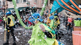 A vibrant street parade scene featuring people in colorful costumes adorned with blue wigs and green cloaks. Confetti fills the air, adding to the festive atmosphere. In the background, spectators line the street, some holding umbrellas, while surrounding buildings provide an urban backdrop.