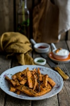 A plate of roasted sweet potato fries garnished with herbs sits on a rustic wooden table. In the background, a cloth napkin and a small dish of sauce are visible along with a fork and a wooden cutting board.