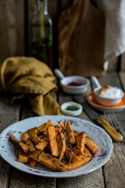 A plate of roasted sweet potato fries garnished with herbs sits on a rustic wooden table. In the background, a cloth napkin and a small dish of sauce are visible along with a fork and a wooden cutting board.