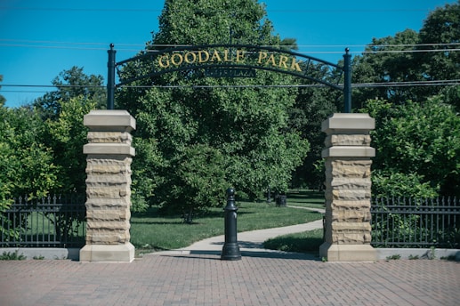 An entrance archway with the words 'Goodale Park' displayed in gold lettering. The arch is supported by two stone pillars, with a grassy park visible in the background. Trees and a paved walkway can be seen beyond the arch.