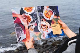 A person holds an open book featuring a vibrant image of a meal with burgers, fries, and drinks. Four people around a table share the food, with the image set against a geometric-patterned tabletop. The book also includes text titled 'Class 8' with language objectives. The background shows a blurred view of the ocean and rocky shoreline.