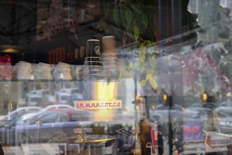 A cafe window display featuring coffee cups stacked on an espresso machine. Reflections of the street outside, including cars and trees, blend with the view inside. There are plants and various kitchen items, and a sign on the machine reads 'La Marzocco.'