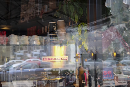 A cafe window display featuring coffee cups stacked on an espresso machine. Reflections of the street outside, including cars and trees, blend with the view inside. There are plants and various kitchen items, and a sign on the machine reads 'La Marzocco.'