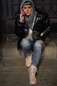 A person with long gray hair, wearing a black puffer jacket, gray hoodie, and jeans, is sitting on stone steps while holding a phone to their ear. They have tan boots and appear to be focused on a conversation. The background includes a detailed iron gate and a dark setting, suggesting an urban environment.
