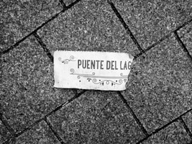 A piece of paper with decorative elements and the words 'Puente del Lago' lies on a textured, gray cobblestone pavement arranged in a diagonal pattern.