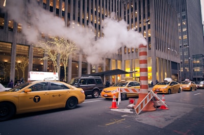 A busy city street scene with several yellow taxis lined up, a tall building with illuminated windows in the background, and a steam pipe emitting a large plume of white steam. The street is adorned with festive lights wrapped around street trees, and traffic cones surround the steam pipe.