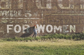 A young girl stands in front of a large brick wall with faded white paint, featuring the words 'FOR WOMEN.' The background mainly consists of the aged, textured brick, and the foreground shows green grass.