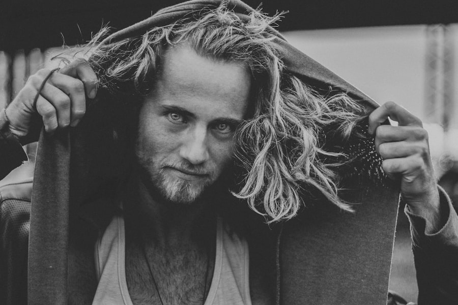 A long-haired man in an undershirt raises a hood over his head and stares at the camera. Photo in greyscale.