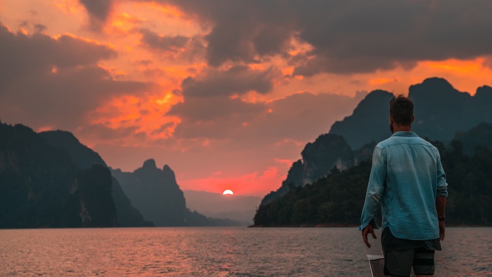 man standing near edge facing body of water during sunset