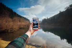 person holding iPhone while taking picture of mountains and body of water during daytime