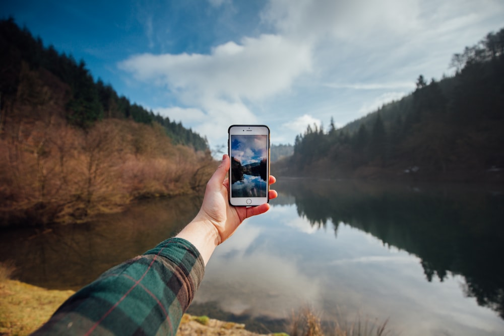 person holding iPhone while taking picture of mountains and body of water during daytime