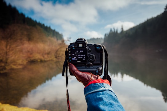 A person wearing a denim jacket with a red glove is holding a DSLR camera, capturing a serene landscape with a calm lake surrounded by trees. The reflection of the surrounding trees and cloudy sky is visible on the lake's surface.