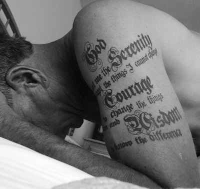 The image features a close-up of a man's shoulder and arm, displaying a prominent tattoo with the words 'God, Serenity, Courage, Wisdom' in stylized lettering.