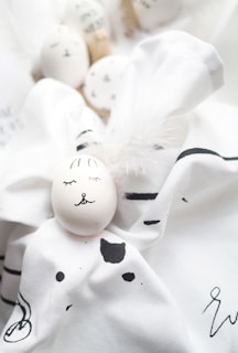 Several white eggs are artistically decorated with simple facial features and placed on a soft, white fabric with black patterns. The arrangement includes fluffy white feathers, contributing to a gentle and whimsical aesthetic.